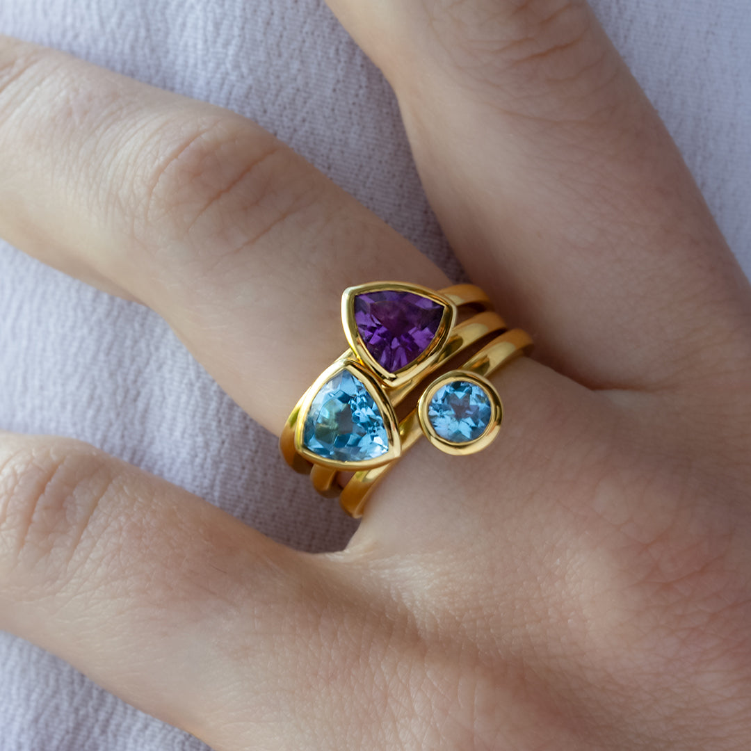 Mano con Combinación de 3 anillos de Oro Vermeil amarillo, con piedras amatista natural tallada en forma triangular y topacio azul natural en forma triangular y brillante.