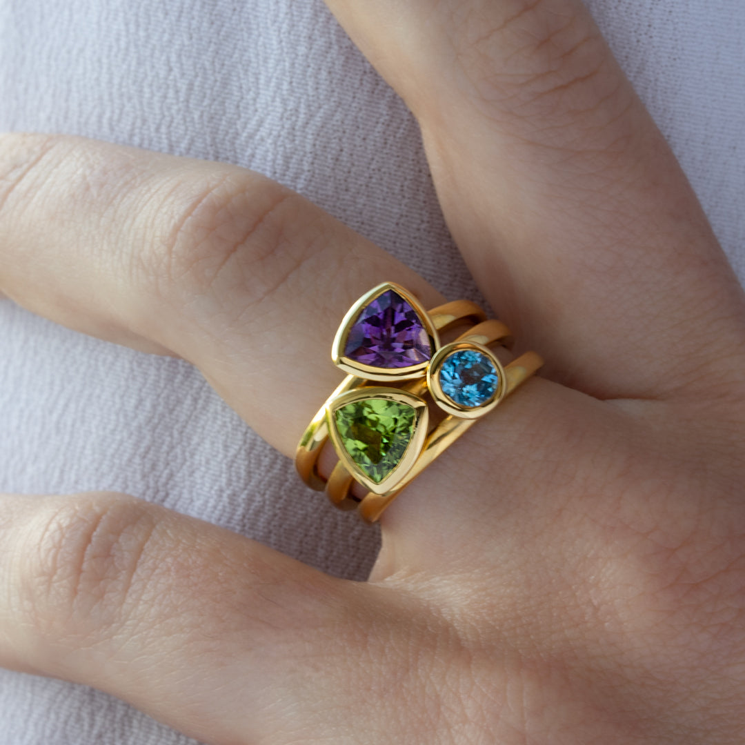 Mano con combinación de 3 anillos de Oro Vermeil amarillo, con piedras amatista natural tallada en forma triangular, peridoto natural en forma triangular y topacio azul brillante.